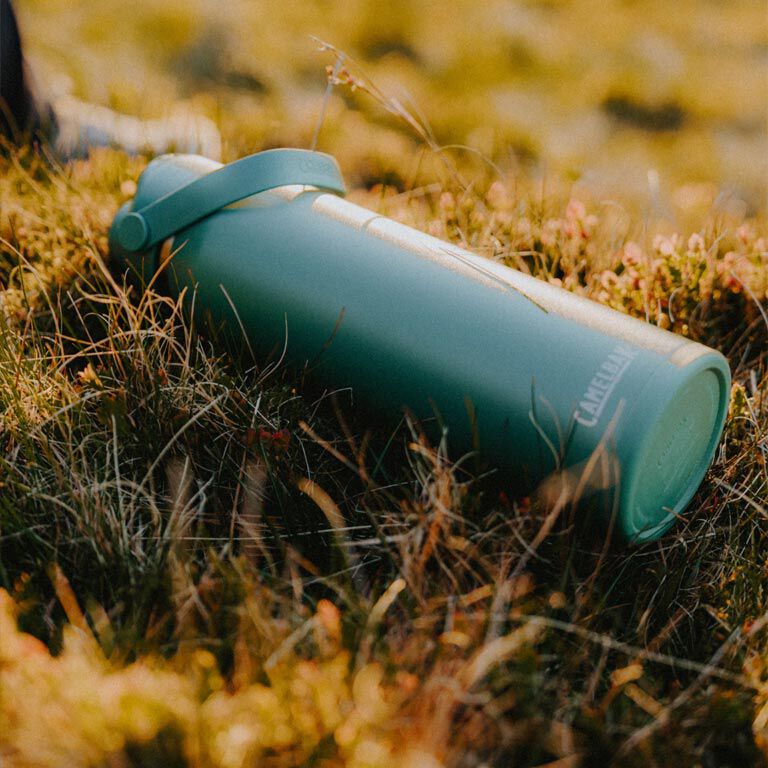 Garrafa Térmica Thrive Camelbak 1,18L repousando sobre a grama em uma paisagem natural.
