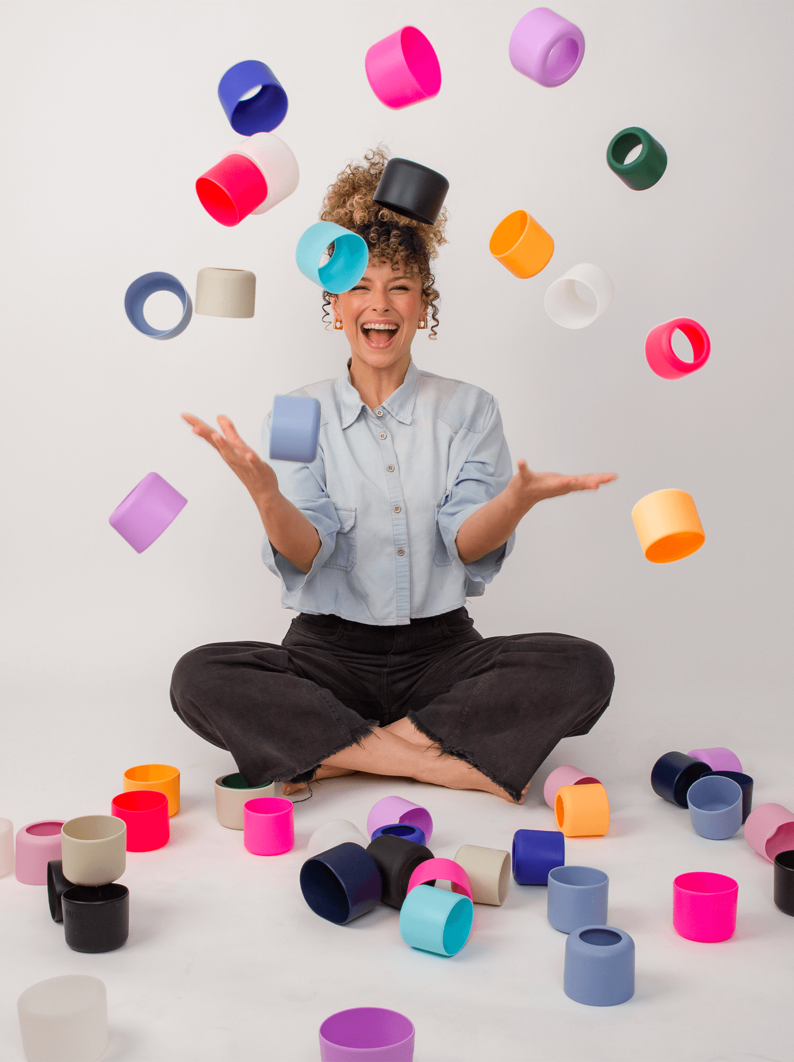 Mulher sorridente brinca com capas coloridas para garrafas VYTA sentada no chão, destacando a versatilidade e diversão das capas PACCO.