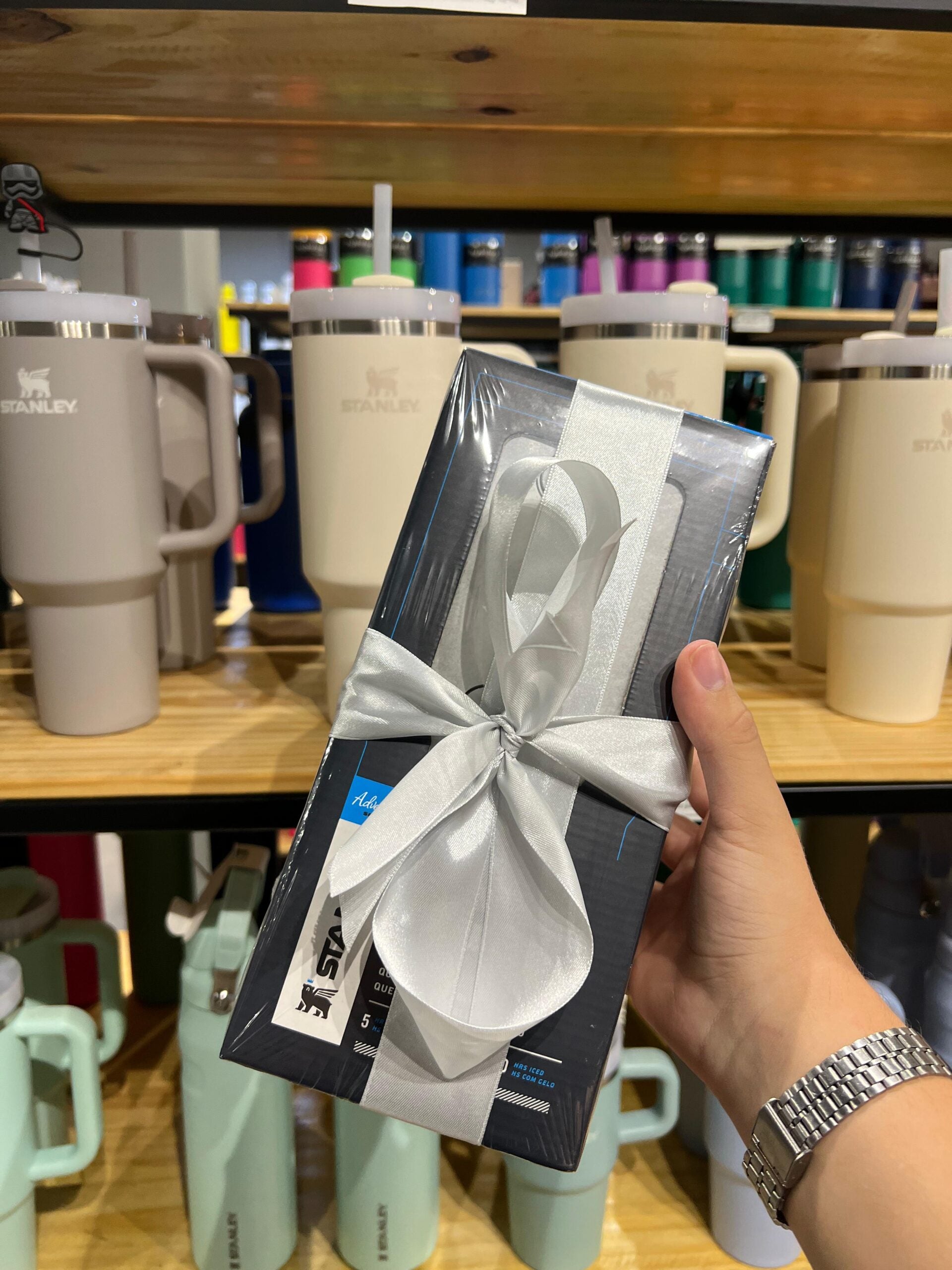 Gift wrapping em uma loja, mostrando embalagem com laço de cetim prateado e termo encolhível, na frente de copos Stanley.