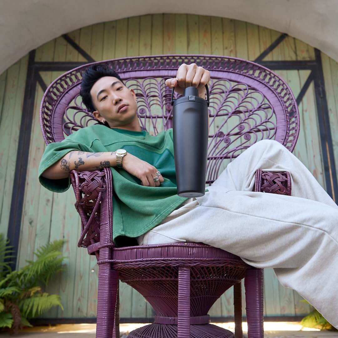 Pessoa relaxando em cadeira roxa, segurando Garrafa Térmica Owala FreeSip Sway - 887ml, destacando estilo e praticidade.