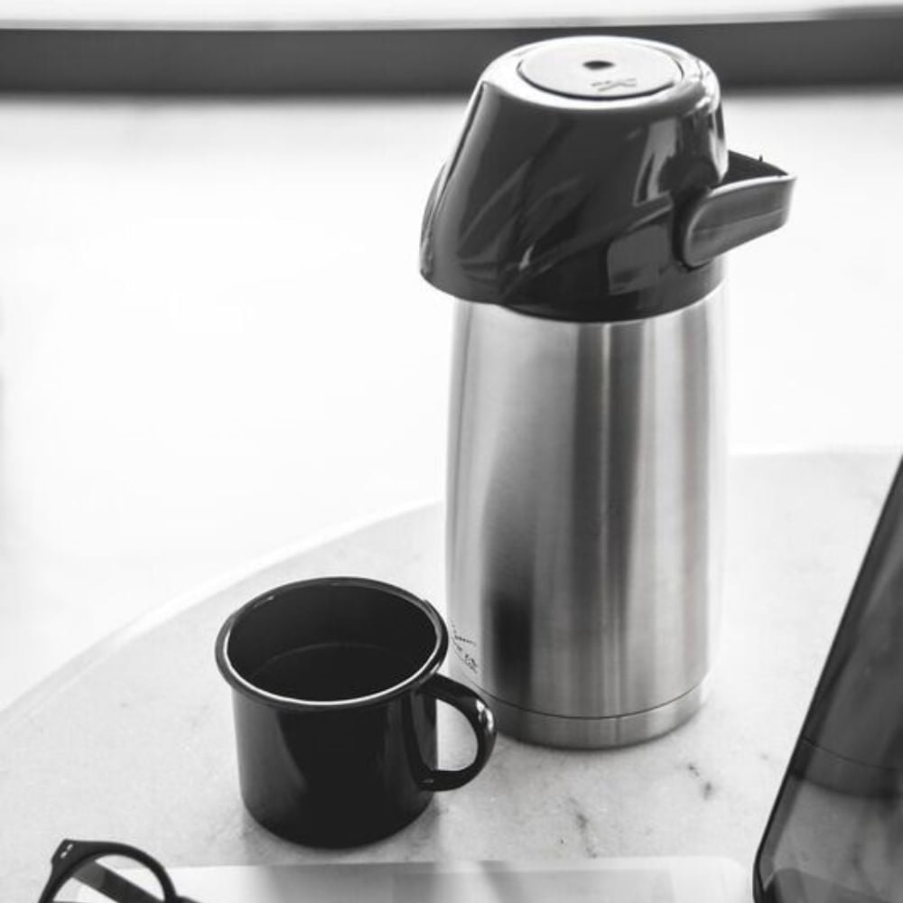 Garrafa Térmica Air Pot Inox Inquebrável 1L Invicta com sistema de pressão e alça integrada ao lado de uma xícara na mesa.