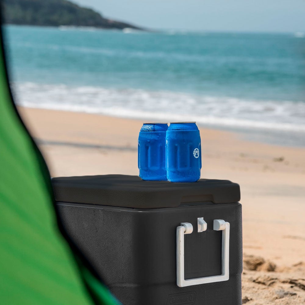 Caixa Térmica 26,5L Personal All Black Coleman na praia com latas azuis sobre a tampa, destacando conservação de alta performance.