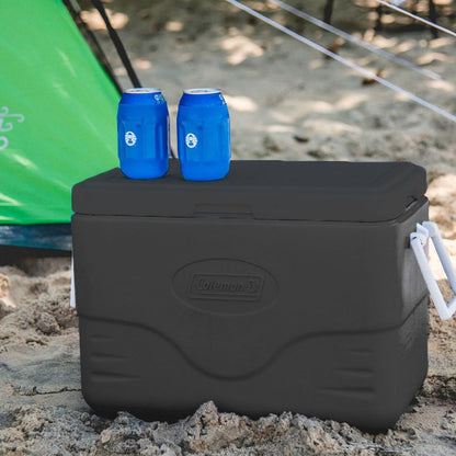 Caixa Térmica 26,5L Personal All Black Coleman na praia com latas de refrigerante em cima, ideal para eventos ao ar livre.
