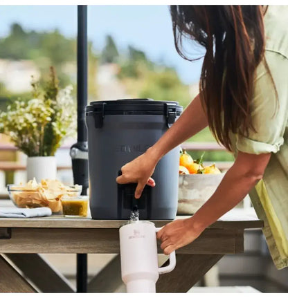 Pessoa usando Jug Térmica Stanley 7,5L em um piquenique ao ar livre, servindo bebida fresca. Ideal para várias ocasiões.