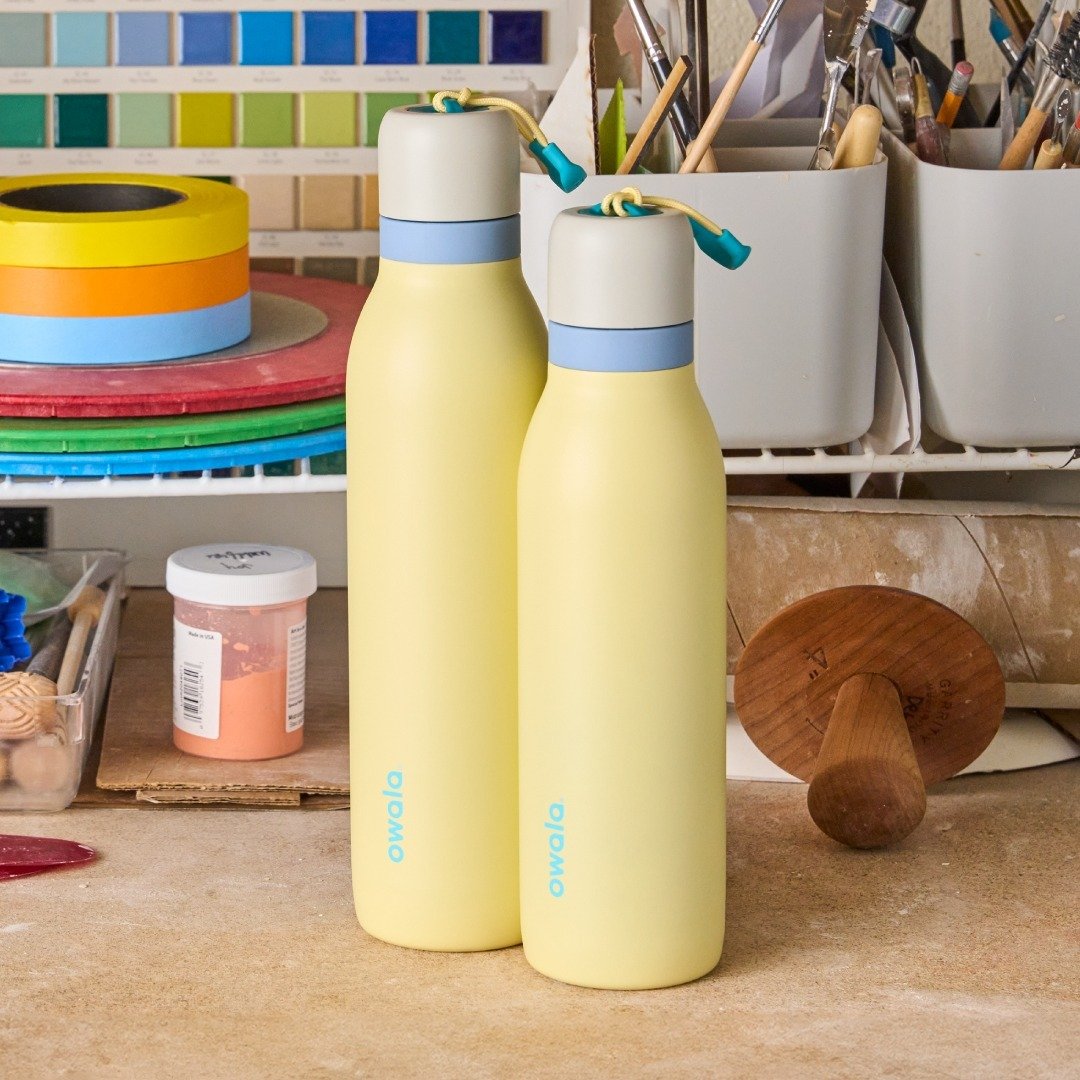 Duas Garrafas Térmicas Owala Twist - 532ml | 18oz em uma mesa de trabalho artística com ferramentas e materiais de arte ao fundo.