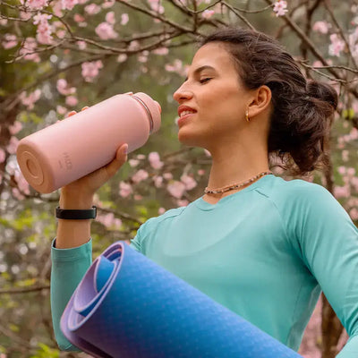 Mulher usando Garrafa Térmica PACCO Hydrav2 950ml, ideal para manter bebidas frias ou quentes, em ambiente natural com flores ao fundo.