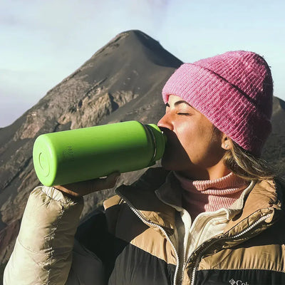 Mulher bebendo de uma Garrafa Térmica PACCO Hydrav2 950ml verde em cenário de montanha, destacando isolamento térmico superior.