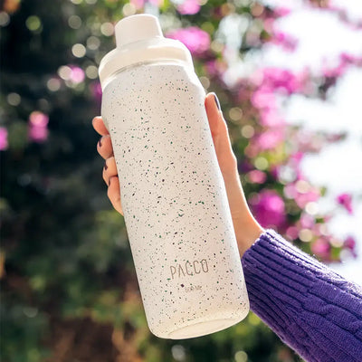 Pessoa segurando Garrafa Térmica PACCO Vyta 500ml, elegante e durável, com fundo desfocado e flores ao fundo.