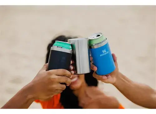 Pessoas brindando com Can Cooler Stanley 296ml na praia, destacando sua eficiência em manter bebidas geladas.