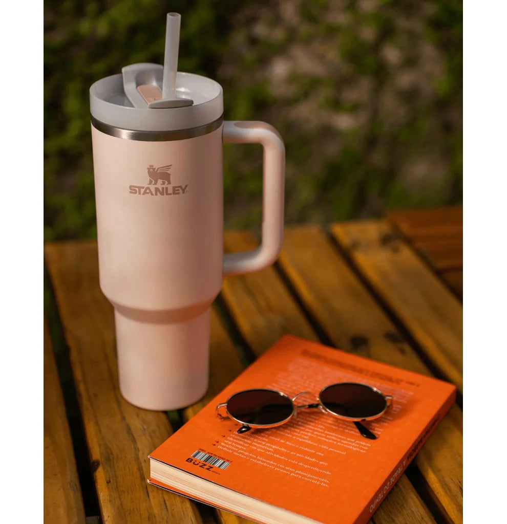Copo Quencher Stanley 1,18L com alça ergonômica em mesa de madeira ao lado de livro e óculos de sol, ideal para hidratação diária.