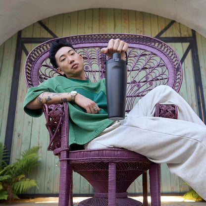 Pessoa relaxando em cadeira roxa, segurando Garrafa Térmica Owala FreeSip Sway - 887ml, destacando estilo e praticidade.