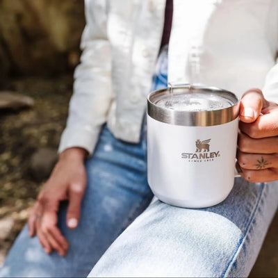 Pessoa segurando Caneca Camp Mug Stanley - 350ml ao ar livre, destacando estilo e durabilidade em um design atemporal.