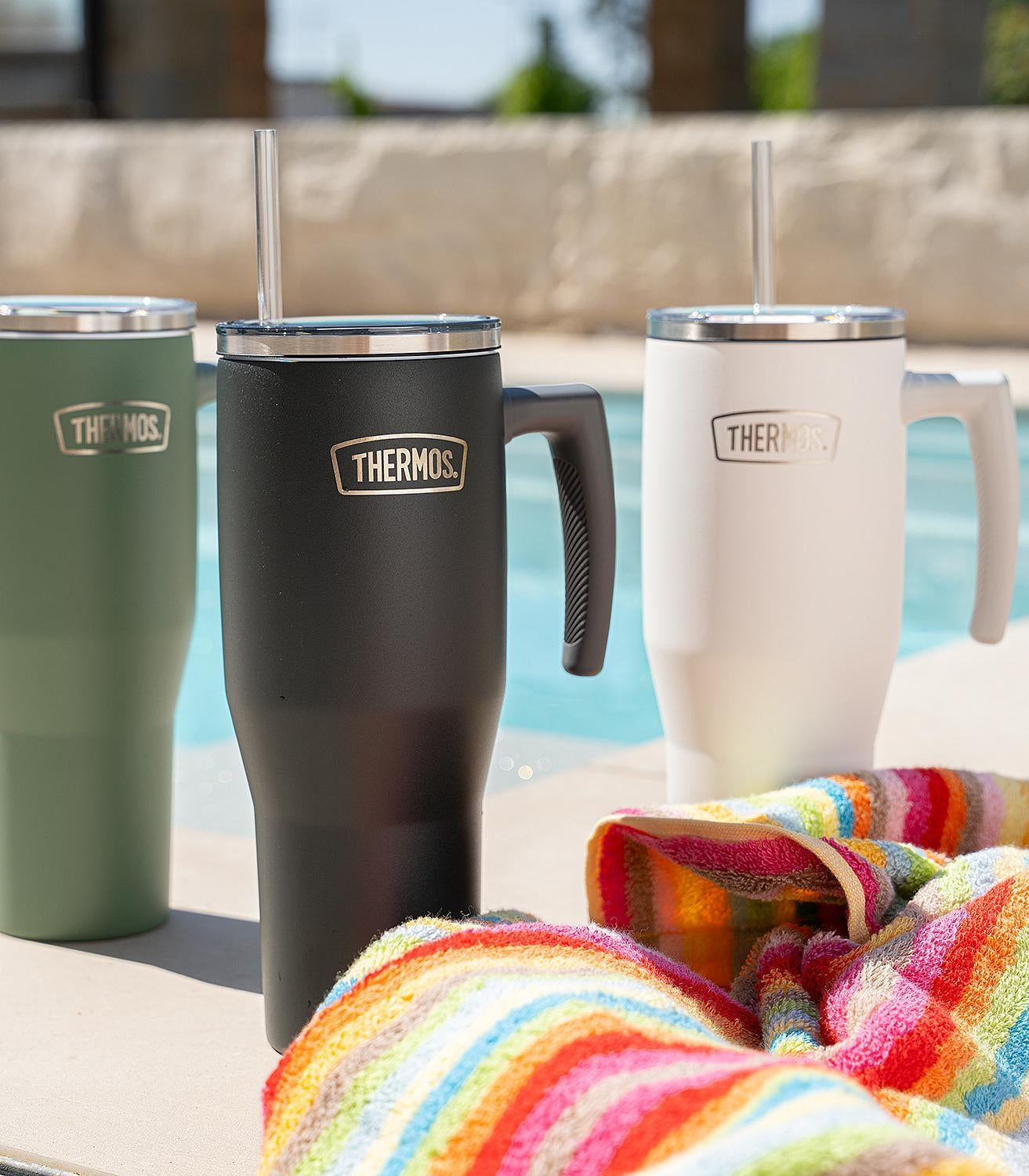 Três copos térmicos Thermos Copo Térmico Aruba com Canudo -1,1L ao lado de toalha colorida, cenário ao ar livre.