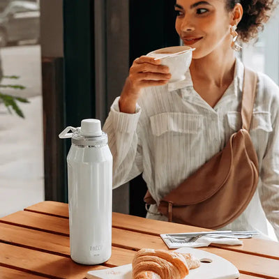 Garrafa Térmica Vyta 950ml PACCO | PEDRAS PRECIOSAS em mesa de café com mulher ao fundo, destacando elegância e eficiência térmica.